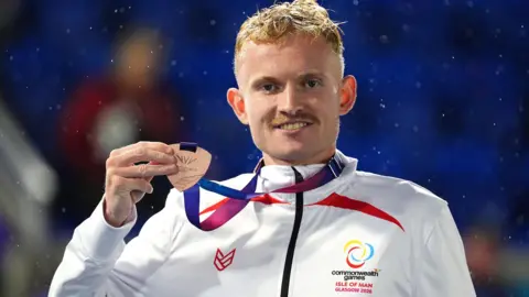PA Mullarkey stood, holding his bronze medal up. His jacket is an Isle of Man team jacket. 