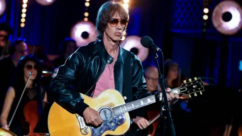 Richard Ashcroft - a man with dark hair, sunglasses and an acoustic guitar, standing at a microphone