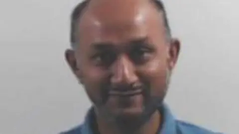 Head-and-shoulders portrait of a person with a closely cropped hairstyle wearing a blue polo shirt, facing the camera against a plain light gray background.