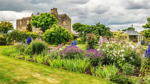 DarloMur A landscaped garden, featuring a bank of flowers and shrubs, including blue lupins or delphiniums, and pale pink roses, behind a grassed area. Behind are some small trees, and to the right a pergola-type structure with pitched roof and open glassed doors, and to the left Thornton Hall - a stone-built 16th century manor house with mullioned windows.