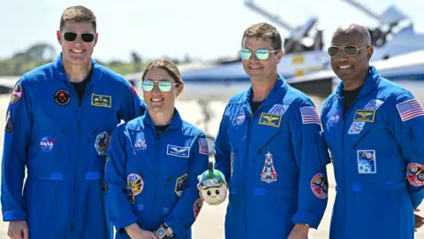 AFP via Getty Images Four Artemis II astronauts stand side by side on a sunny runway, posing for a group portrait. They all wear bright blue NASA flight suits covered in mission patches and name badges, with dark boots. One astronaut in the centre holds a small mascot or model in both hands. Behind them, two sleek white-and-blue T‑38 training jets sit on the concrete, their pointed noses facing left and right, with the NASA “meatball” logo visible on a tail fin. The sky above is clear and pale blue, giving the scene a crisp, formal but upbeat feel.