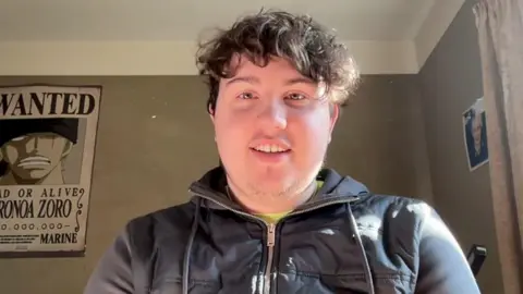 BBC Fintan Hood, a young boy with curly brown hair, smiling in his room at the University of York. He is wearing a black zipped up jacket.