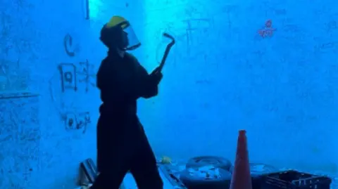 BBC Picture of someone in a blue light-lit rage room surrounded by items to smash. They are wearing a yellow helmet and holding a crowbar. Among the items are a crate, car tyres and a traffic cone. There is rudimentary graffiti on the walls.