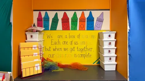A brightly coloured classroom display shows large cartoon crayons with smiling faces above a yellow sign that reads, “We are a box of crayons. Each one of us unique, but when we get together, our classroom is complete.” On the left are stacked trays and folders in a red rack, and on the right is a tower of white storage boxes. Colourful sheer fabrics are spread out on the floor in front of the display.