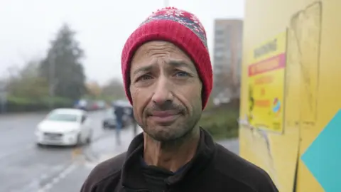 Andy wearing a red bobble hat and black jumper. He is looking at the camera without much expression on his face. He is stood next to yellow boarding and has a car going past him to the left of the image.