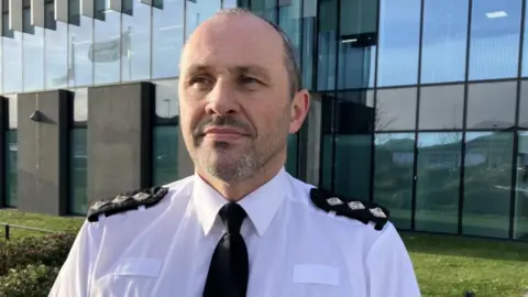 Ch Insp Stuart McIver has short dark hair and a short beard. He is wearing a white police uniform shirt and black tie and is standing in front of a large glass-fronted building.