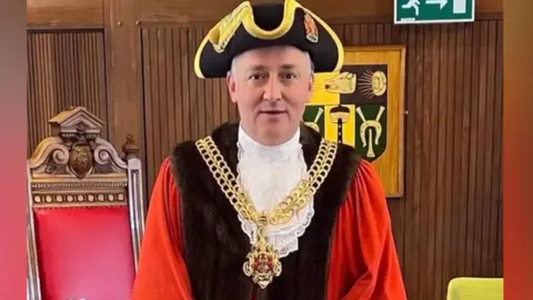 A false image of Ashley Bowkett the mayor of Gloucester in his mayoral robes ceremonial chain and traditional hat. The fake image depicts him standing in the council chamber with a large old wooden traditional chair behind him with a red leather back and a wooden panelled wall. 
