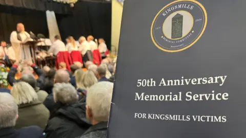 A board that says Kingsmills Memorial Committee. 50th anniversary memorial service for Kingsmills victims. A crowd is watching a minister speak on a stage. 
