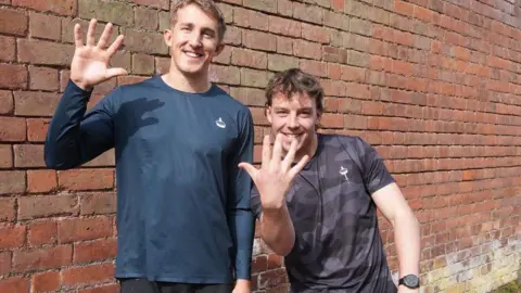 Alfie (Left) and Joe Pritchard (Right). Both men are raising their hands to the camera and the taller man on the left is wearing a blue top and has blonde hair. The man on the left has brown hair and they are both smiling. 