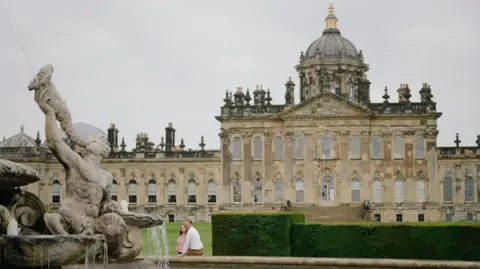 Castle Howard. A large stately home. A couple sit on the edge of a fountain in front of the property. 