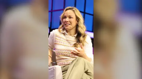 Eline Van der Velden has long blonde hair. She is seated, and wearing a white shirt, sleeveless jumper and beige trousers.