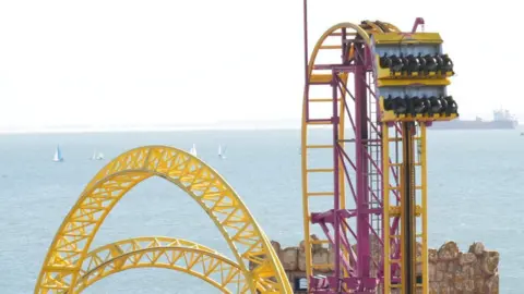 Richard Knights/BBC A yellow rollercoaster cart, with two rows containing four black seats each, nears a high point in the track. A large ship and smaller sailing boats can be seen on the water in the background