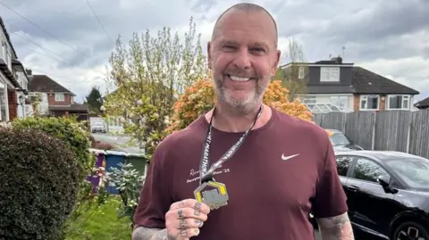 Grant Williams Grant Williams has short shaved hair and salt and pepper beard. He has a maroon Nike T-shirt and is holding his Newport Marathon medal around his neck.