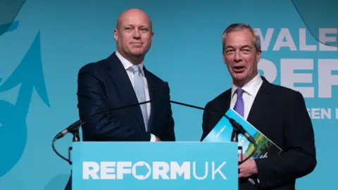 Dan Thomas and Nigel Farage, men in suits, holding Reform manifesto at manifesto launch in Wales for the Senedd elections