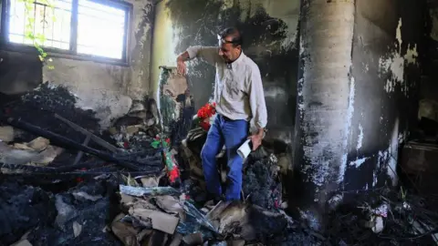 'Israeli forces took over my home and then they set it on fire' BBC A man in a pale shirt and jeans stands in a room blackened by fire holding burnt items including colourful artificial flowers.