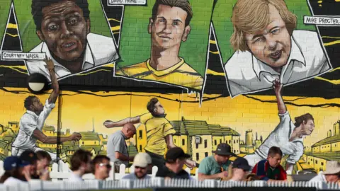 Getty Images A group of cricket fans sit watching a match in Bristol under a large mural showing three former Gloucestershire players. From the left are painted Courtney Walsh, David Payne and Mike Procter.