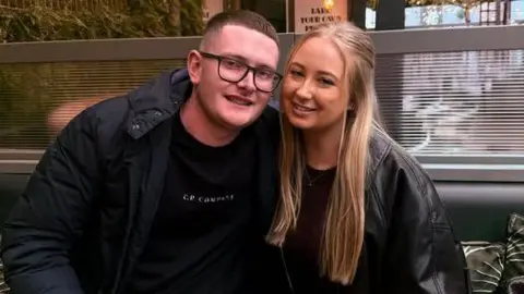 Facebook Jamie Fountain has short cropped brown hair and is wearing black rimmed glasses, a black padded jacket and a black T-shirt. He is sitting next to Ellie Ireland, who has blonde long hair and is wearing a dark leather jacket and a black top.