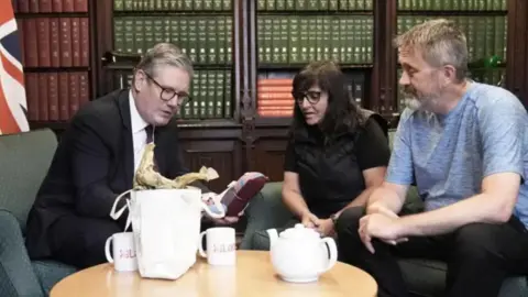 PA Media Prime Minister Keir Starmer (left) with Figen Murray (centre) and her husband Stuart sitting in a room with cabinets containing books