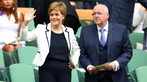 Getty Images A woman with short hair and a white jacket is smiling and waving. Next to her is a bold man in a smart blue suit and white shirt, his cufflinks are visible. The picture is taken in a sports stadium. 