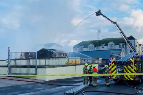 Fire engine and firefighters, with plume of grey smoke, and a height vehicle being used to dampen down the scene, with football stadium floodlights in the background.