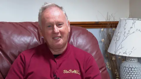 Huw Samuel, wearing a red top sitting on a red leather seat beside a lamp and smiling