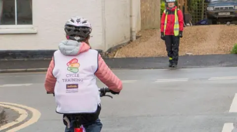 A child wearing a pink puffer coat, silver crash helmet and a white high-vis jacket rides on a bike along a road. An adult wearing a silver helmet, red coat and yellow high-vis jacket is stood on a pavement ahead of the child.