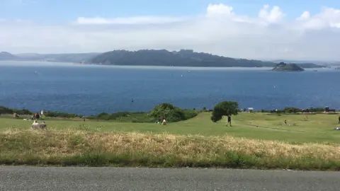 A view from a car park across a green cliff in front of blue sea and showing an island and Cornwall in the distance
