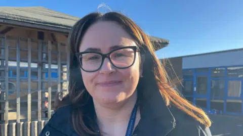 Kizzie pictured looking into the camera and smiling. She's wearing a black winter coat with a blue Windmill Primary lanyard just visible. She's stood in a school playground with a large wooden pagoda-type structure to her right. There's school buildings with large glass doors behind her to her left. The sky is bright blue and clear, with lots of sunshine. 