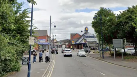 Google Bilston High Street with shops, a road with double yellow lines and cars driving on it, lampposts and pedestrians walking nearby.