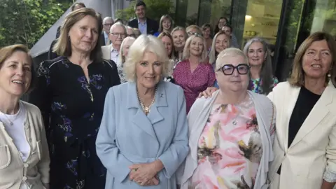 PA Media Queen Camilla wearing a blue dress stands in the centre of five women smiling at the camera. Men and women stand behind looking towards the camera.