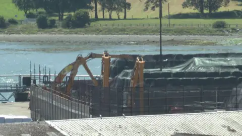 A screengrab of footage of Warrenpoint Port, zoomed in on a close-up of Re-Gen's waste storage facility. Rows of bales of compacted black bin waste, wrapped in black plastic-like covers, are lined up on the harbour. Several large yellow mechanical diggers are beside the bales. The roof of a port building is partially visible in the foreground and the blue waters of Carlingford Lough and its opposite shore are in the background.