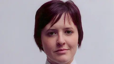 Philippa Reynolds in a police photo. She has short purple/red hair and is wearing her uniform. She is standing in front of a white wall. 