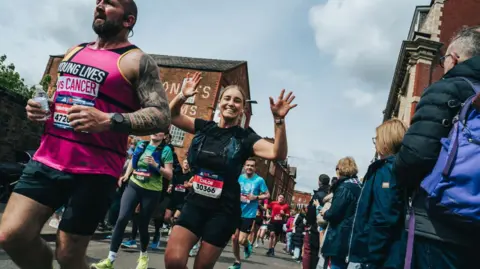 A runner in the Manchester Marathon smiles and waves her hands in the air as she and hundreds of others jog along a road. Spectators line the side of the road.