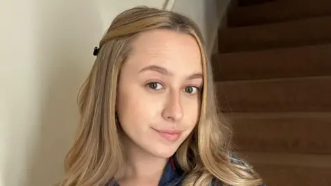 Cait Yardy A selfie of Cait with blonde hair with wooden stairs behind her