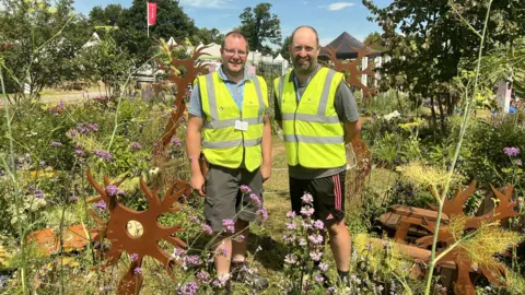 Two men wearing hi-vis stand in the garden they've created with many flowers and sculptures at the Badminton Flower Show