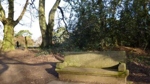 Shotover Country Park, with a memorial seat made of stone in the foreground, a dirt path stretching between some trees, and a dogwalker in the distance