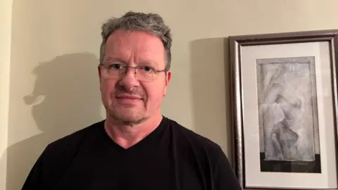 Martin Heath/BBC John Cook with short grey hair and glasses, wearing a black v-neck sweater and smiling at the camera. There is a framed modern picture to his left and he is standing in front of a cream-coloured wall.