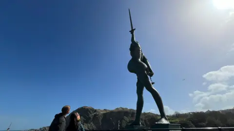 A large statue of a human figure stands on a seafront, holding a long sword pointed up into the sky, with steep green cliffs rising behind it. In the foreground, two people stand side by side looking up at the statue under a clear blue sky with the sun shining brightly.
