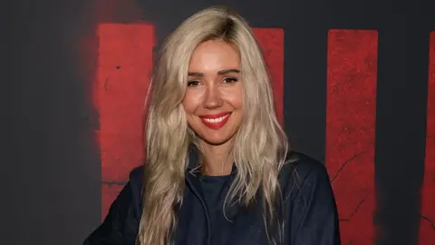 Getty Images Alanah has long blonde hair and is wearing a navy blue jacket and top. She has red lipstick on and stands in front of a black background with red writing on it 