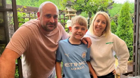 James smiles at the camera, with his parents on either side of him. Mike wears a light pink shirt, while James wears a blue Manchester City shirt and Hannah wears a very light grey hoodie.