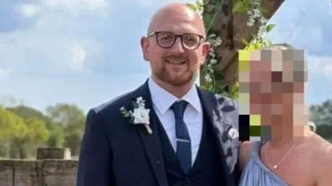 Facebook Gary Carson, who has a bald head, ginger beard and glasses, smiles at the camera wearing a navy blue suit, waistcoat and blue tie over a white shirt. He is posing with his arm around a woman in a blue dress whose face has been blurred out. 