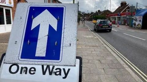 BBC A blue one way sign on the pavement next to a road lined with shops