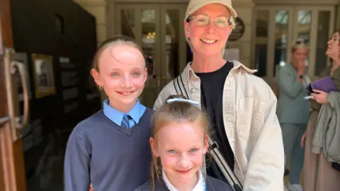 A woman, wearing glasses, and a baseball cap, and a cream coloured denim jacket is standing with two young girls. Both girls have light brown hair tied away from their faces. The taller one at the back is wearing a grey v necked jumper over a pale blue shirt. The smaller one at the front of the picture is wearing a grey cardigan over a blue gingham dress. They are all smiling.