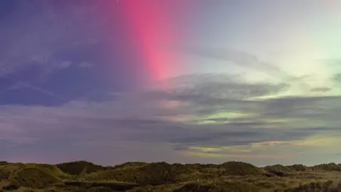 Christian Spooner Red, pink and green hues in the sky, dotted with cloud, above a landcape of grass covered sand dunes .