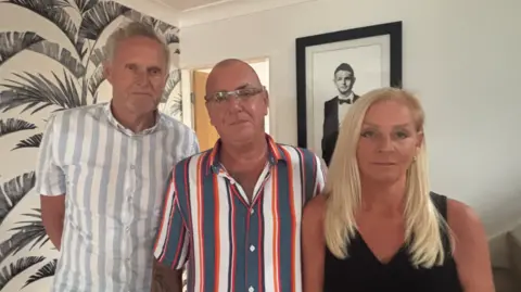 A man with grey hair and a grey and white striped shirt next to a bald man with glasses and a red white and blue striped shirt and a woman with long blonde hair and a black top in a white-walled room