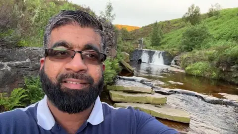 Faisal Mahmood A man with black greying hair and a black beard takes a selfie in front of a waterfall. He wears varifocal glasses and a blue polo shirt with a white collar.