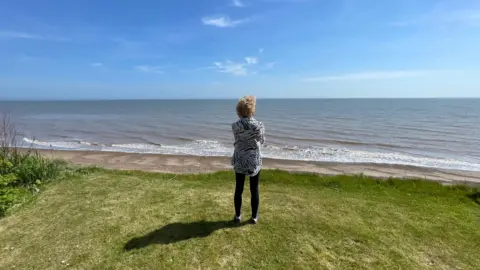 A woman is standing on grass at the top of a cliff and is looking out to sea