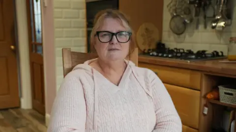 Shelley Herniman is a middle-aged woman, wearing a pink jumper and black-rimmed spectacles, sitting on a chair in a kitchen.