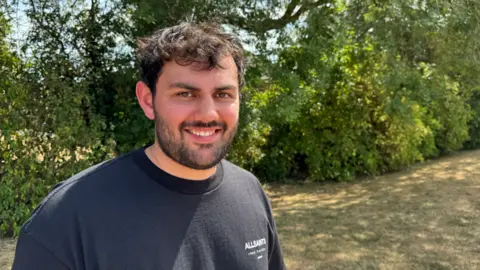 A man in his 20s is smiling. He has black hair and a black beard and is wearing a black t-shirt. He is standing in a park.