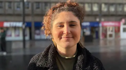 Image of a young woman looking into the camera. She has tied back hair. She is standing on a shopping street and is smiling.